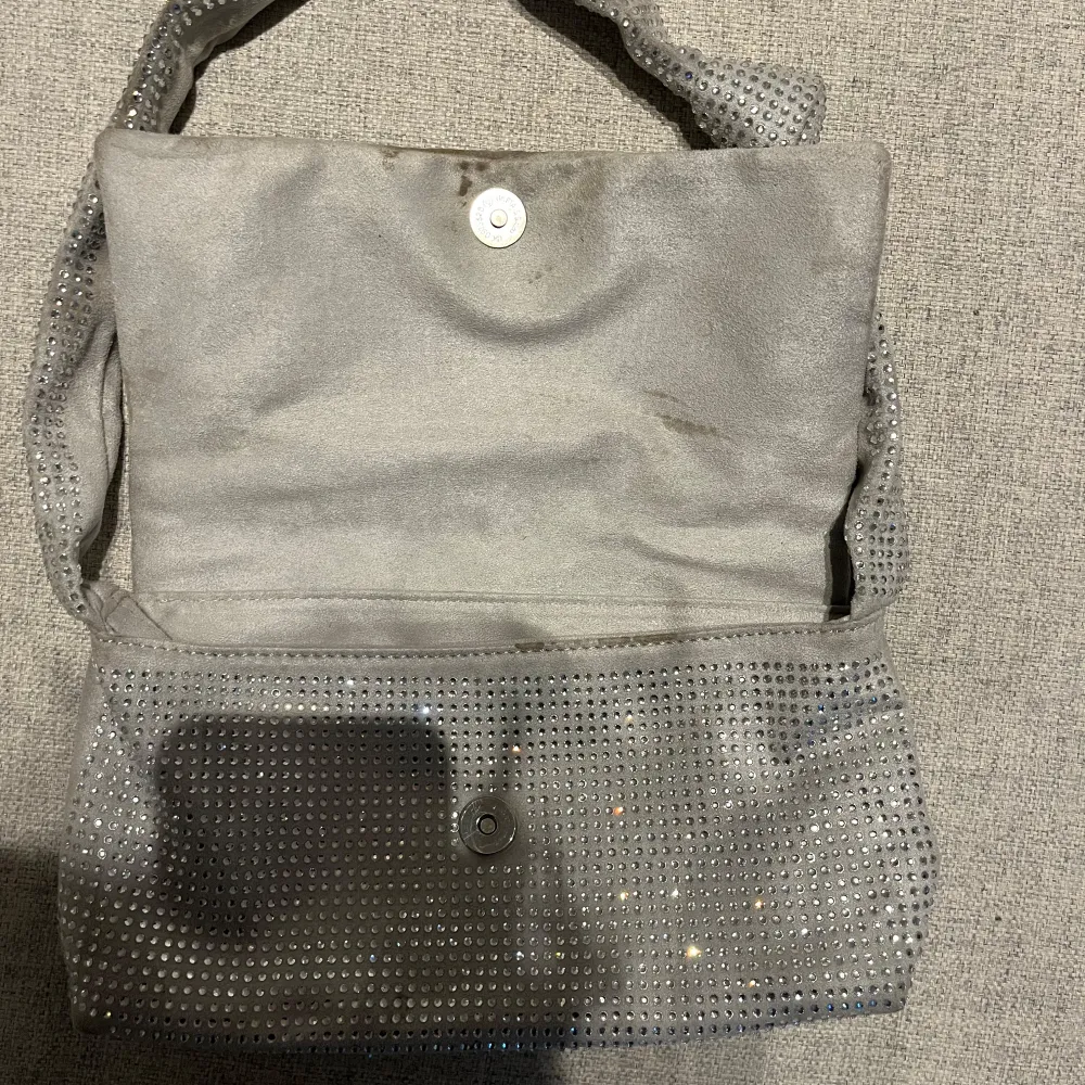 Snygg grå axelväska med glittriga detaljer från Gina tricot. Väskan har en praktisk magnetknäppning och ett innerfack med dragkedja för extra säkerhet. Perfekt för att ge din outfit lite extra bling!. Laukut & Käsilaukut.