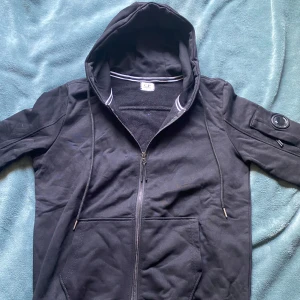 Svart hoodie från C.P. Company - Säljer en svart hoodie från C.P. Company med dragkedja och huva. Hoodien har en logga på ärmen och snören vid huvan. Storlek S men skulle säga att den passar som XS också. Endast använd några gånger. Pris kan diskuteras vid snabb affär!