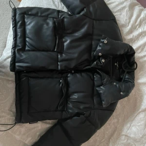 Svart pufferjacka från Missguided - Säljer en snygg svart pufferjacka från Missguided. Jackan har en hög krage och stora fickor framtill. Perfekt för kyliga dagar med sin vadderade design. Den har långa ärmar och stängs med dragkedja och knappar. Ett måste för garderoben!