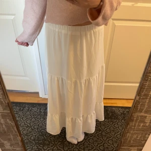 Vit långkjol med volang - Säljer en stilren vit långkjol med volangdetaljer och en slits nertil. Kjolen har en elastisk midja för bekväm passform. Perfekt för sommaren och matchar med vad som helst. Den har en underkjol vilket gör att den ej är genomskinlig!🩷