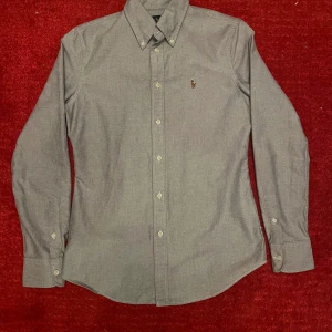 Ralph lauren skjorta  - Säljer denna snygga gråa skjortan från Ralph lauren. Den är i mycket bra skick utan defekter. Skjortan sitter slimfit alltså lite tajtare längs kroppen