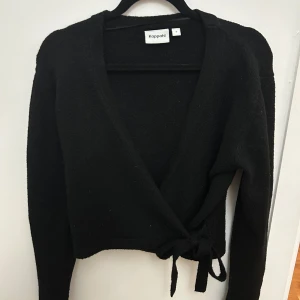 Svart omlottopp från Kappahl - Snygg svart omlottopp från Kappahl med knytning i sidan. Toppen är croppad och har en v-ringad design som ger en stilren look. Perfekt för att skapa en trendig outfit.