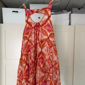 Färgglad midi klänning med mönster - Säljer en färgglad midiklänning med ett unikt mönster i rosa, orange och vitt. Klänningen har en luftig passform och är ärmlös, perfekt för varma sommardagar. Materialet är lätt och fladdrigt, vilket ger en skön känsla.