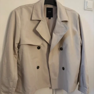Beige trenchcoat - Snygg beige trenchcoat med dubbelknäppning och klassisk krage. Perfekt för vår och höst. Jackan har en stilren design med långa ärmar och en lös passform. Storlek S