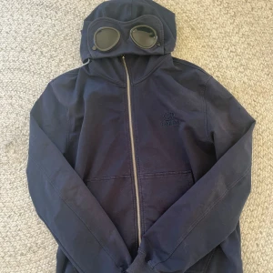 Mörkblå huvtröja från C.P. Company - Säljer en mörkblå huvtröja från C.P. Company med dragkedja och unika glasögondetaljer på huvan. Perfekt för en stilren och avslappnad look. Tröjan har långa ärmar och en praktisk ficka framtill. Ny skick skriv om ni är intresserade 