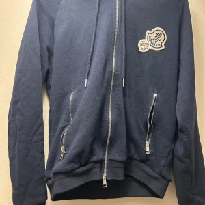 Mörkblå hoodie från Moncler - Säljer en snygg mörkblå hoodie från Moncler med dragkedja och broderad logga på bröstet. Den har långa ärmar och praktiska fickor med dragkedjor. Perfekt för en stilren och bekväm look. Är M men passar mer S