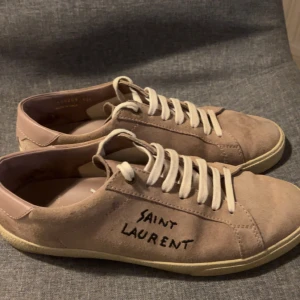 Beige sneakers från Saint Laurent - Snygga beige sneakers från Saint Laurent med klassisk snörning och stilren design. Tillverkade i mocka med märkets logga broderad på sidan. Perfekta för en avslappnad och trendig look.