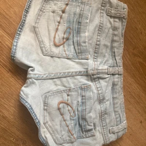Ljusblå vintage jeanshorts med söta detaljer - Snygga ljusblå jeansshorts med coola slitningar och en unik spetsblomma på framsidan. Shortsen har broderade detaljer och en låg midja för en avslappnad look. Perfekta för varma sommardagar!