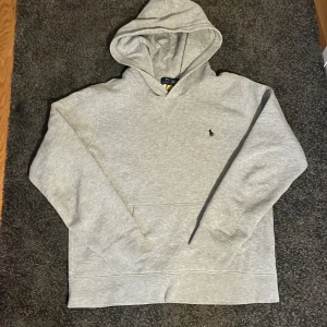 Grå hoodie från Ralph Lauren - Säljer en stilren grå hoodie från Ralph Lauren med den klassiska broderade loggan på bröstet. Tröjan har en bekväm passform med långa ärmar och en praktisk magficka. Perfekt för en avslappnad stil. Tröjan är 160 i barnstorlek.