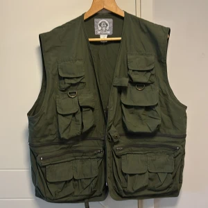 Grön väst från Uncle Milty (Fiskeväst) - Säljer en grön väst från Uncle Milty med flera praktiska fickor och dragkedja. Västen är tillverkad i en blandning av bomull och polyester, vilket gör den både slitstark och bekväm. Perfekt för utomhusaktiviteter och äventyr.