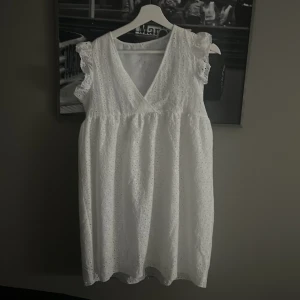 Vit ärmlös klänning med spetsdetaljer - Säljer en söt vit klänning med v-ringning och spetsdetaljer. Klänningen är ärmlös och har en luftig passform, perfekt för varma dagar. Den har en romantisk känsla med sina volangdetaljer vid axlarna. Studentklänning. 