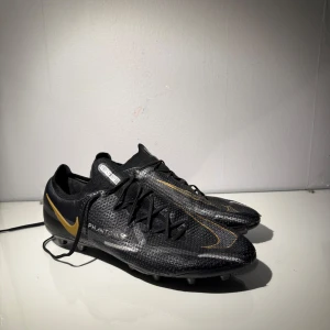 Nike PHANTOM  - Snygga svarta Nike fotbollsskor med gulddetaljer och snörning. Skorna har en modern design med en diskret Nike-logga i guld på sidan. Perfekta för fotbollsplanen med en tight passform för optimal kontroll.