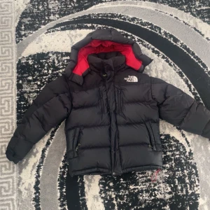 Svart dunjacka från The North Face - Säljer en svart dunjacka från The North Face med röd insida. Jackan har en huva och dragkedja framtill samt två fickor med dragkedjor. Perfekt för kalla dagar.