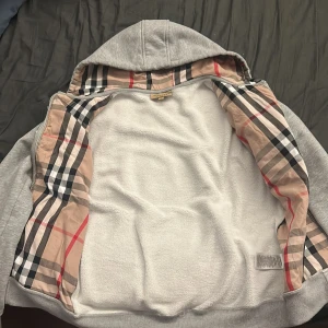 Grå hoodie från Burberry - Säljer en stilren grå hoodie från Burberry med klassiskt rutigt mönster på insidan av huvan. Den har dragkedja framtill och praktiska fickor. Perfekt för en avslappnad look nu i våren och sommaren. Burberry påse ingår.