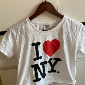 Vit I Love NY t-shirt - Säljer en klassisk vit t-shirt med det ikoniska 'I Love NY'-trycket i svart och rött. T-shirten är kortärmad och tillverkad i 100% bomull, vilket gör den bekväm och lätt att bära. Perfekt för alla som älskar New York! 🌆 Den visar lite mage men super fint till sommaren