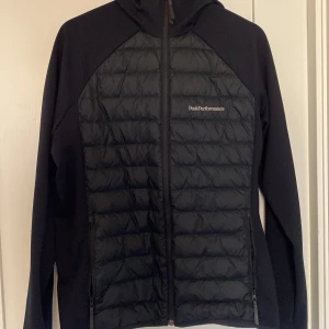 Svart jacka från Peak Performance - Säljer en svart jacka från Peak Performance med dragkedja och huva. Jackan har en quiltad design på framsidan och baksidan, vilket ger extra värme och stil. Perfekt för kyligare dagar.