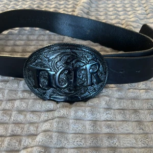 Tiger of sweden - Snyggt svart bälte med ett stort, detaljerat spänne där det står 'TIGER'. Perfekt för att ge en cool touch till din outfit. Bältet är gjort av skinn och har en robust känsla.