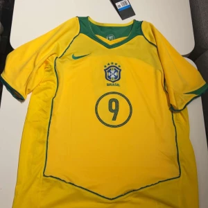  Brasilien Hemmaställ 2004 - Säljer en gul Brasilien fotbollströja från Nike med gröna detaljer och nummer 9 på både fram- och baksidan. Tröjan har korta ärmar och är perfekt för fotbollsfans. Den har även texten 'Ronaldo' på baksidan.