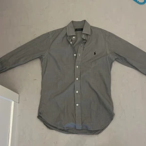 Grå skjorta från Ralph Lauren - Snygg grå skjorta från Ralph Lauren med klassisk krage och knappar framtill. Skjortan har långa ärmar och en broderad logotyp på bröstet. Perfekt för en stilren look.