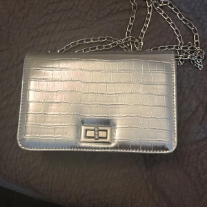 Silverfärgad axelväska med krokodilmönster - Snygg silverfärgad axelväska med krokodilmönster och kedjerem. Väskan har en elegant låsdetalj framtill och en stilren design som passar perfekt för en kväll ute.