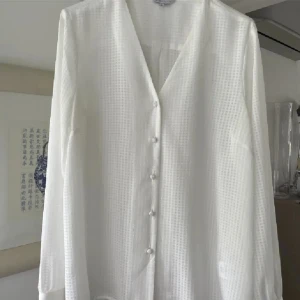 Vit blus från Style Sixty Five - Elegant vit blus från Style Sixty Five med ett subtilt rutmönster. Blusen har en v-ringning och knappar framtill. Perfekt för en stilren look. Mycket fint skick eller aldrig använd, kommer inte ihåg. 