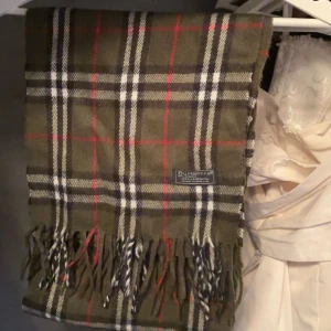 Rutig halsduk från Burberry - Snygg grön rutig halsduk från Burberry med inslag av rött, svart och vitt. Den har klassiska fransar i ändarna och är perfekt för att hålla dig varm och stilren under kyliga dagar.