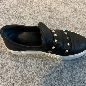 Svarta skor med nitar - Snygga svarta sneakers i skinn med coola nitar på ovansidan. De har en vit sula som ger en skön kontrast och en stilren look. Perfekta för att ge din outfit en touch. Skorna är i använt skick. 