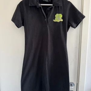 Svart klänning från 2117 of Sweden - Snygg svart klänning från 2117 of Sweden  med korta ärmar och krage. Den har en grön detalj framtill och siffran '03' i grönt på baksidan. (Katt och kanin finns i hemmet)