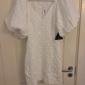 Vit klänning från Gina Tricot - Säljer en elegant vit klänning från Gina Tricot med puffiga ärmar och v-ringning. Klänningen har en vacker brokadstruktur och en dragkedja i ryggen. Perfekt för speciella tillfällen, exempelvis studenten🤍
