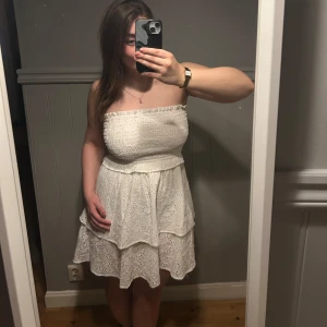 Vit off shoulder klänning - Säljer en söt vit axelbandslös klänning med volanger och broderade detaljer. Perfekt för sommarens alla tillfällen. Klänningen har en smockad överdel som ger en bekväm passform.