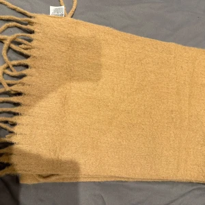 Beige halsduk från H&M - Säljer en stilren beige halsduk från H&M. Den är mjuk och har fransar i ändarna som ger en extra touch. Mått: 200 cm x 43 cm.