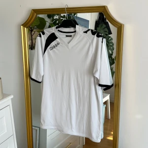 Vit tränings tröja från Kappa - Säljer en vit fotbollströja från Kappa med svarta detaljer på axlarna och ärmsluten. Tröjan har en V-ringad hals och korta ärmar. Perfekt för träning eller match! Trycken på axlarna har slitage