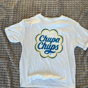 Vit Chupa Chups t-shirt - Säljer en vit t-shirt med Chupa Chups-logga i blått och gult. T-shirten har en normal passform och korta ärmar. Perfekt för en avslappnad stil!