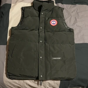Svart dunväst från Canada Goose - Säljer en svart dunväst från Canada Goose med en klassisk design och varumärkets logotyp på bröstet. Västen har knappar framtill och två stora fickor. Perfekt för kyliga och sommar dagar. Den är aldrig använd, den är helt ny. Pris kan diskuteras 
