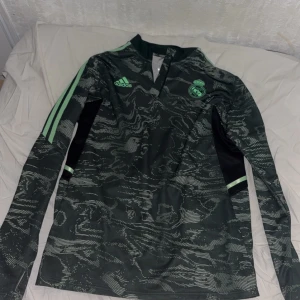 Real Madrid tröja - Snygg långärmad fotbollströja från Adidas i mörkgrön med ljusgröna detaljer och ett unikt mönster. Tröjan har klubbens emblem på bröstet och de klassiska Adidas-ränderna på ärmarna. Perfekt för fotbollsfans som vill sticka ut på planen.