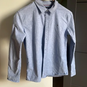Ljusblå skjorta från Tommy Hilfiger - Snygg ljusblå skjorta från Tommy Hilfiger med klassisk krage och diskret logga på bröstet. Skjortan har långa ärmar, knappar framtill och är gjord i mjuk bomull. Perfekt för dig som gillar stilrena och tidlösa plagg.