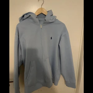 Ljusblå hoodie från Ralph Lauren - Säljer en ljusblå hoodie från Ralph Lauren i storlek M men lite mindre i storlek. Tröjan har huva, fickor framtill och är tillverkad i mjuk bomull. Perfekt för chill dagar eller när du vill ha en clean look. Mycket bra skick förutom ett litet hål längst ner på ärmen men inget som syns