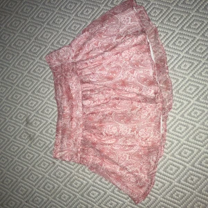 Rosa mönstrad kortkjol från Gina Tricot - Supersöt kortkjol från Gina Tricot i rosa och vitt med paisleyinspirerat mönster. Kjolen har resår i midjan och är gjord i ett lätt och luftigt material, perfekt för varma dagar. Passar dig som gillar en feminin och trendig stil.