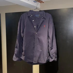 Mörkblå skjorta från Ralph Lauren - Snygg mörkblå skjorta från Ralph Lauren i slim fit-modell. Skjortan har klassisk krage, långa ärmar och knappar framtill. Ikoniska röda loggan broderad på bröstet. Perfekt för dig som gillar stilrena och tidlösa plagg.