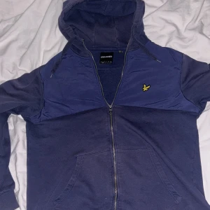 Blå hoodiejacka från Lyle & Scott - Mörkblå hoodiejacka från Lyle & Scott med dragkedja och huva. Jackan har en gul logga på bröstet och praktiska fickor framtill. Materialmix med bomull och polyester, perfekt för chill dagar. Snygg och enkel stil som funkar året runt.