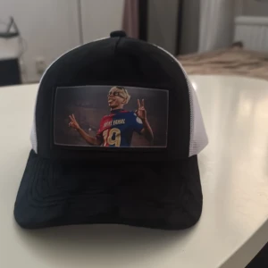 Svartvit keps med Lamine Yamal-tryck - Cool svart och vit truckerkeps med mesh baktill och justerbar passform. Framtill finns ett stort tryck av fotbollsspelaren Lamine Yamal i matchtröja. Perfekt för dig som gillar fotboll och vill sticka ut med en unik accessoar.