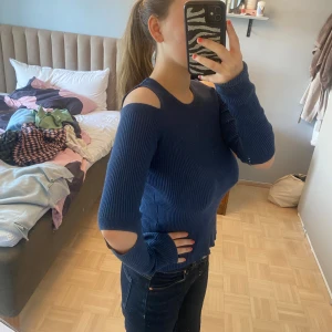 Festtopp med öppna axlar och armar  - Säljer en mörkblå ribbstickad tröja med coola cut-outs på axlar och armar. Tröjan har lång ärm och sitter figurnära. Perfekt för dig som vill ha en unik och trendig look. Passar till jeans eller kjol för en snygg outfit.