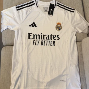 Real Madrid vit matchtröja Adidas - Snygg vit Real Madrid fotbollströja från Adidas med svarta detaljer och klubbmärke på bröstet. Tröjan har korta ärmar, svarta ränder på axlarna och är tillverkad i lätt, ventilerande material med AEROREADY-teknologi. Perfekt för dig som älskar fotboll och vill bära klubbens färger.
