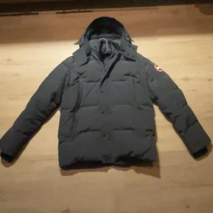 Canada Goose Wyndham grå - Säljer en svart dunjacka från Canada Goose i storlek M. Jackan har huva, klassisk logga på ärmen och en clean, puffig look. Perfekt för kalla dagar med sin rejäla fyllning och snygga passform. Flera fickor framtill och diskreta knappar.