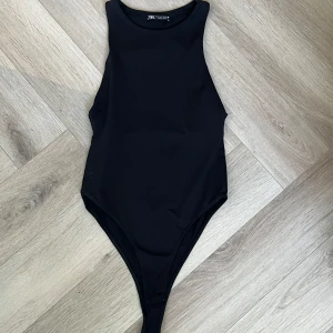 Svart ärmlös bodysuit från Zara - Snygg svart bodysuit från Zara med hög hals och ärmlös design. Tight passform som sitter perfekt till höga jeans eller kjol. Enkel och stilren look, tillverkad i stretchigt syntetmaterial. Knäppning i grenen för smidig användning.