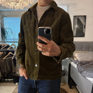 Olivgrön manchester overshirt H&M - Säljer en olivgrön overshirt från H&M i manchester. Jackan har klassisk krage, bröstficka och knäppning framtill med svarta knappar. Perfekt för lager-på-lager och ger en chill vibe till din outfit.