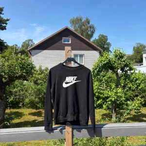 Svart Nike sweatshirt med logga - Svart sweatshirt från Nike med stor vit logga framtill. Klassisk rund halsringning och långa ärmar. Tröjan är i mjuk bomullsmix och har en avslappnad passform som funkar till allt. 