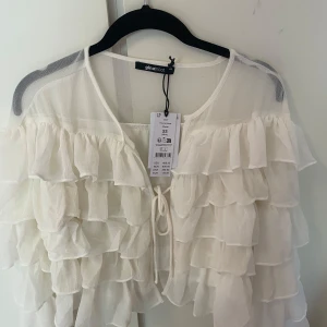 Offwhite volangblus från Gina Tricot - Supersöt offwhite blus från Gina Tricot med flera lager volanger och transparent tyg. Blusen har knytning framtill och långa ärmar, perfekt för dig som gillar romantisk och luftig stil. Passar till både jeans och kjol för en trendig look.