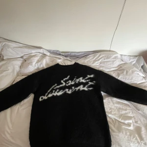 Svart Saint Laurent stickad tröja - Säljer en svart stickad tröja från Saint Laurent med vit, suddig logga framtill. Tröjan har lång ärm, rund halsringning och är gjord i ett fluffigt, mjukt material som känns lyxigt och cozy. Perfekt för dig som vill ha en statement piece med designer-vibe.