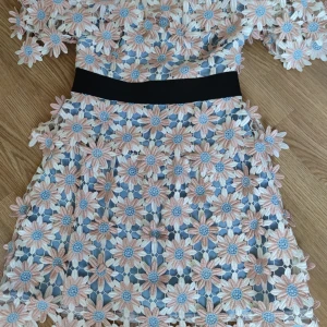 Blommig kortklänning från Self-Portrait - Superfin kortklänning från Self-Portrait med 3D-blommor i pastellrosa, vitt och ljusblått. Klänningen har markerad midja med svart bälte, halvtransparent blommig spets och korta ärmar. Perfekt för dig som vill sticka ut med en unik look!